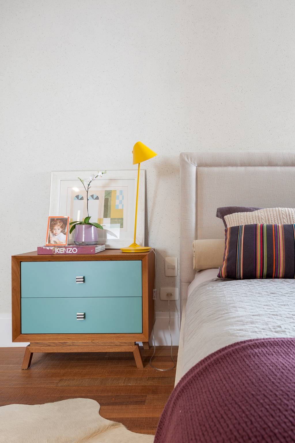 quarto No canto esquerdo, aparece uma mesa de cabeceira de madeira com as gavetas azuis. Sobre ela, há uma luminária de mesa amarela, alguns porta-retratos e um quadro apoiado na parede. Ao lado, aparece metade da cama, que tem cabeceira estofada, almofadas listradas de laranja e marrom e a colcha branca. No pé da cama, há uma colcha roxa