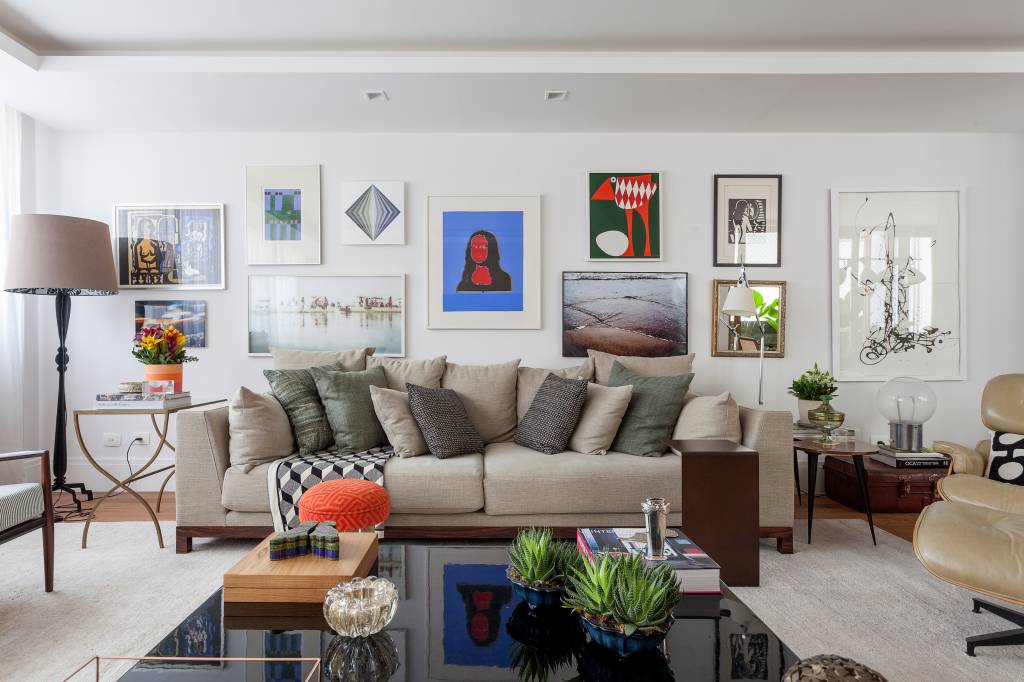 sala-de-estar-gallery-wall Em primeiro plano aparece uma mesa de centro de laca preta bastante grande com vasos e objetos decorativos sobre ela. Ao fundo, um sofá de linho cru com almofadas verdes e uma parede estilo gallery wall, com diversos quadros