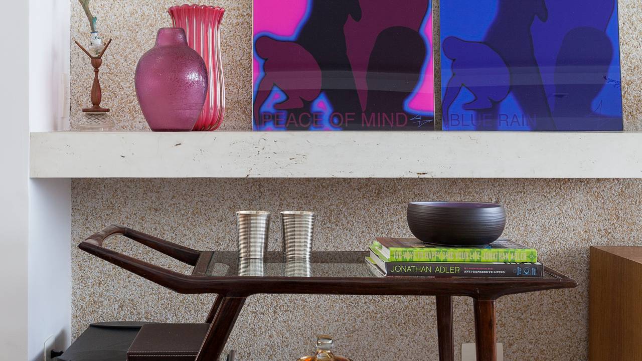 Em um ambiente claro, um carrinho de bar de madeira tem copos, uma cerâmica e algumas embalagens de vidro. Ao fundo, em uma prateleira, há dois quadros, um rosa e um azul, e alguns vasos cor de rosa