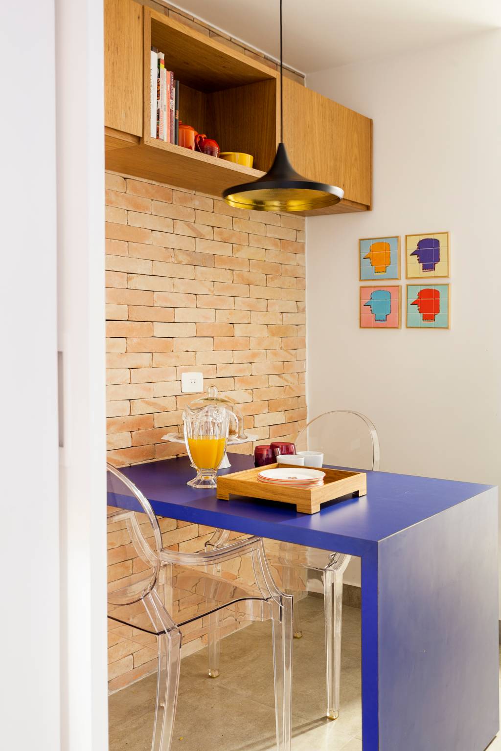 cozinha Na foto, em diagonal, é retratada uma cozinha, com mesa azul e cadeiras transparente. A parede ao fundo é de tijolos aparentes. Há um quadro colorido na paredes. O pendente preto e dourado aparece sobre a mesa, que tem uma bandeja de madeira e jarra de vidro com suco de laranja.