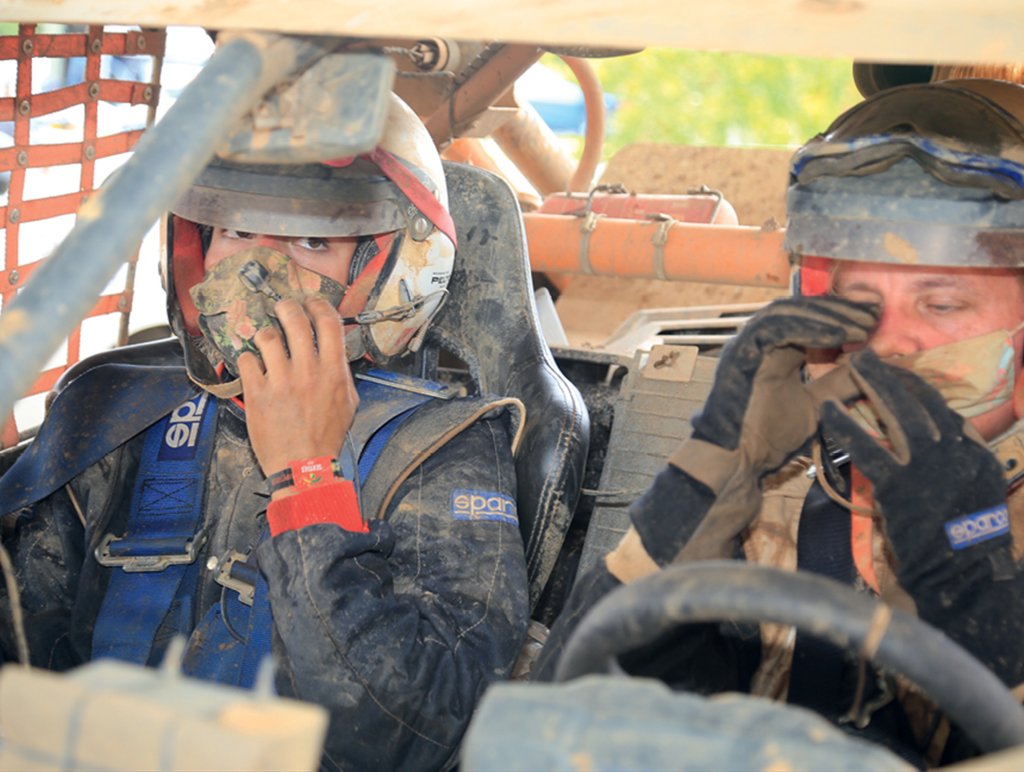 Duas mulheres estão sentadas nos bancos da frente do veículo. Elas estão com o uniforme, máscara e capacete, então só é possível enxergar os olhos delas. Elas e a superfície do carro estão sujas de terra e poeira levantadas na corrida.