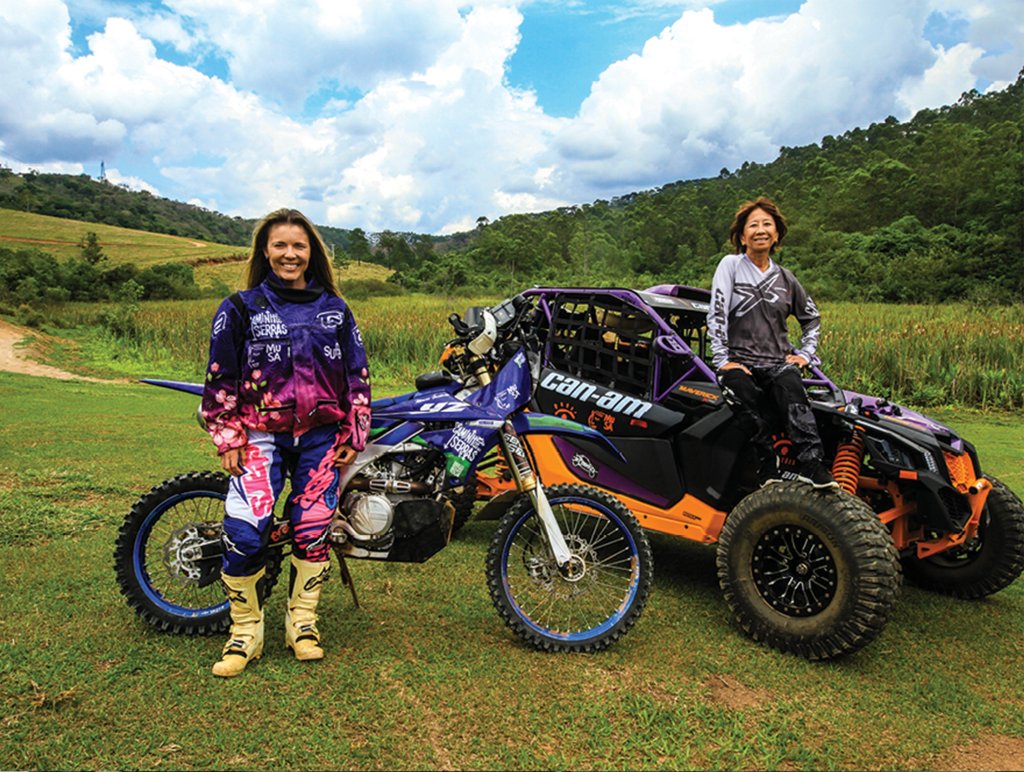 Em um campo verde, duas mulheres vestidas com uniformes estão paradas junto aos seus veículos. Uma delas, loira, tem uma moto, usa uniforme rosa e roxo e botas amarelas. A outra, de cabelos castanhos curtos, usa uniforme cinza e preto e está sentada sobre um UTV