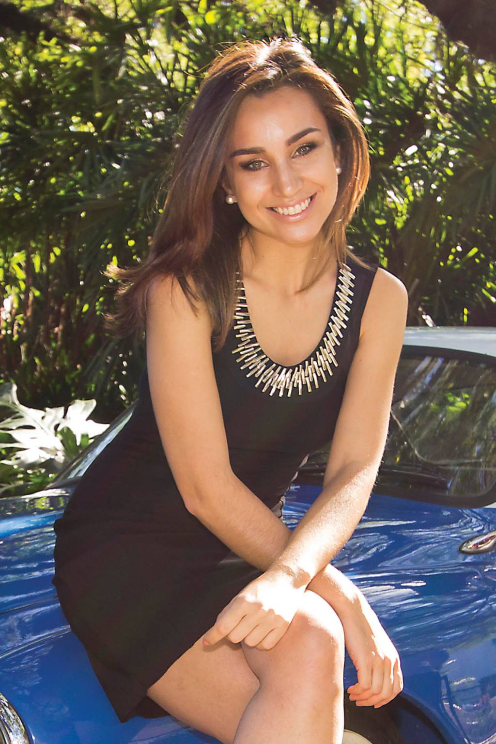Voluntária Millena Machado Vacina Covid Mulher posa para a foto sentada no capô de um carro vintage azul. Ela usa um vestido preto com detalhes prateados na gola e está com as pernas cruzadas, levemente inclinada para o lado.