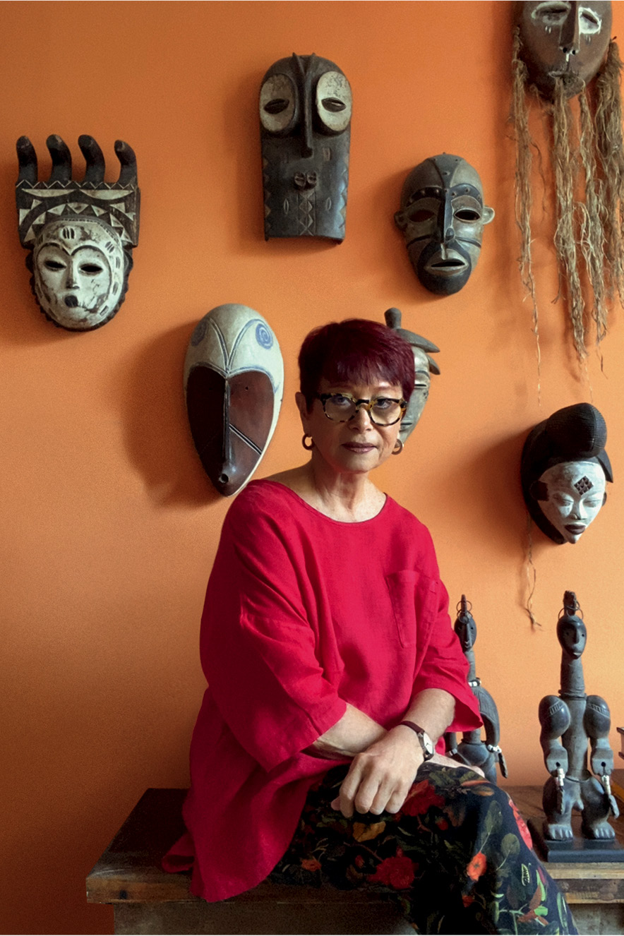 Na sala de sua casa, Julita olha para a c&acirc;mera. Atr&aacute;s dela h&aacute; uma parede com m&aacute;scaras africanas