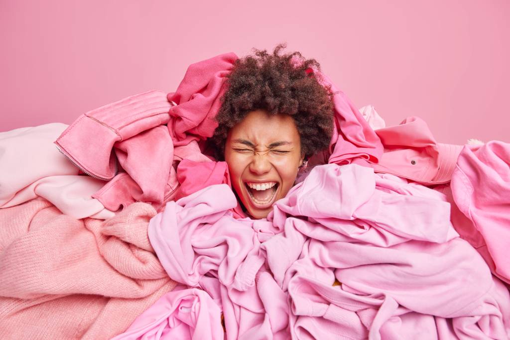 Emotional curly haired woman surrounded by pile of messy clothes from closet exclaims loudly keeps mouth opened has real chaos at home busy doing laundry. Everything in pink color. Clothing concept Roupas
