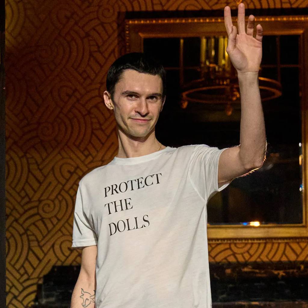 Conner-Ives-proteja-as-bonecas Homem visto de frente, com o braço esquerdo levantado e a mão mostrando dois dedos.
