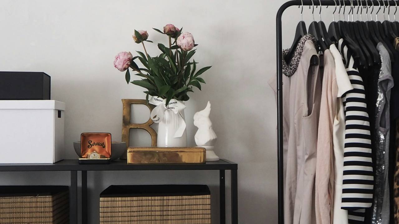 Cabideiro preto com roupas de diferentes cores. Ao lado, mesa preta com jarro de flores e objetos.