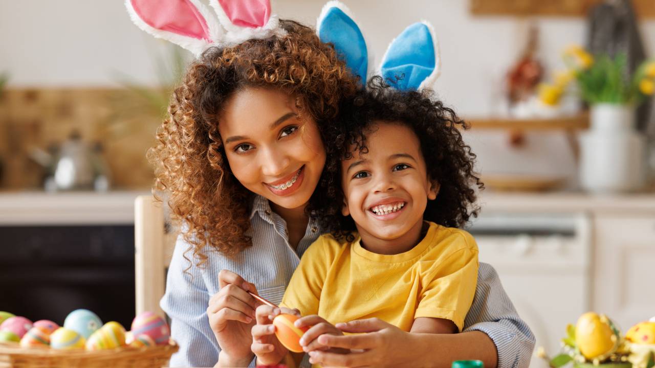Mãe e filho sorridentes na cozinha, preparando sobremesas de Páscoa. Eles usam orelhas de coelho.
