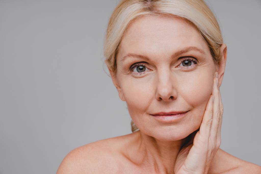 ativos-skincare-menopausa Mulher madura com expressão serena tocando o rosto, em ambiente iluminado naturalmente, representando o autocuidado e a beleza real na maturidade.