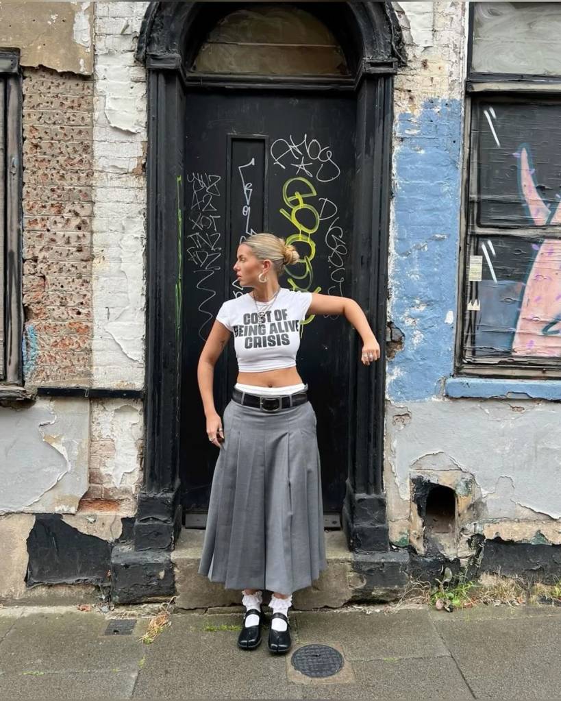 Pessoa com camiseta cropped estampada, saia cinza pregueada, meia branca com babado e sapato tabi branco. Visual jovem e lúdico.