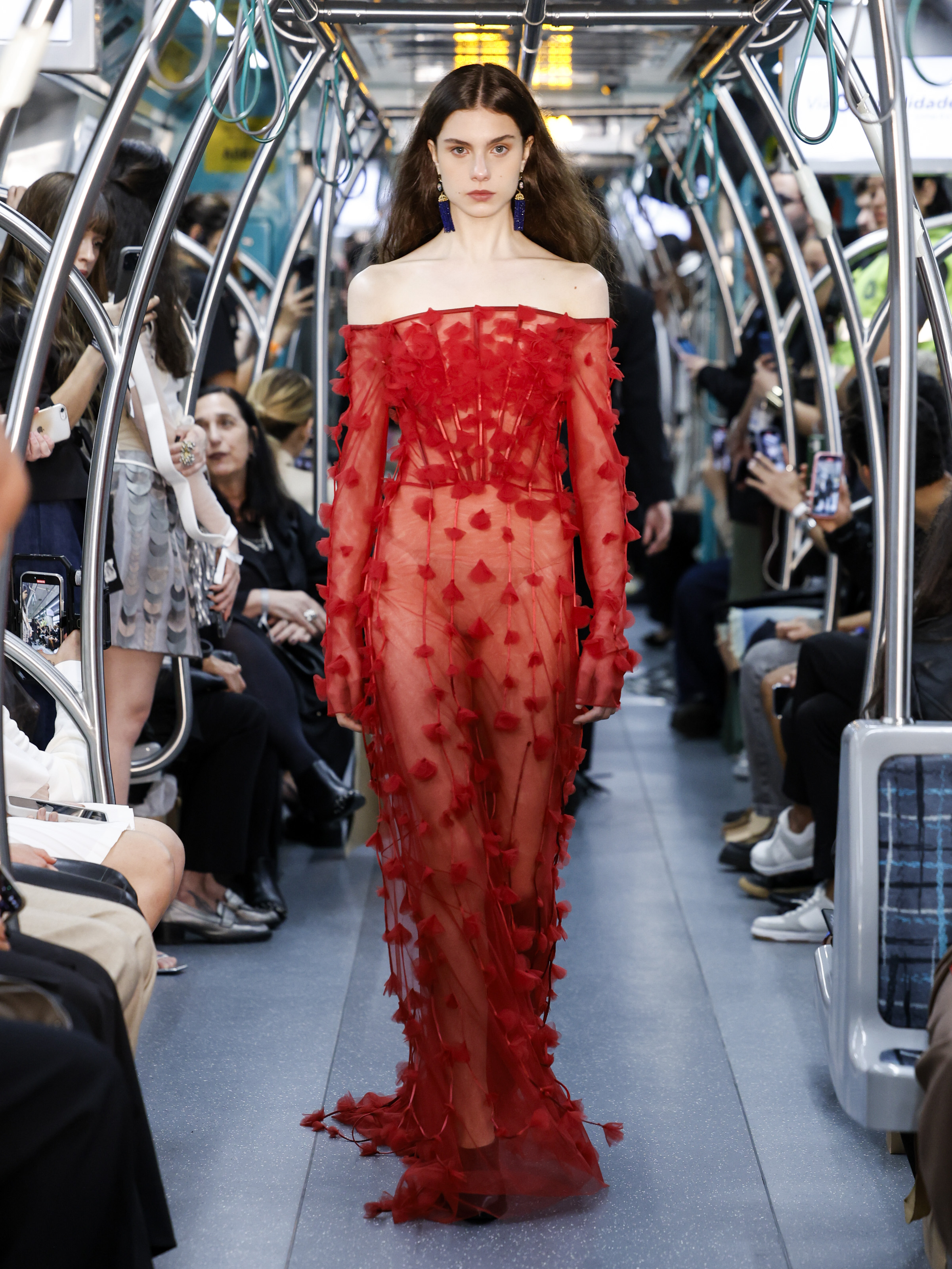 Foto de uma modelo desfilando na SPFW. Ela usa um vestido vermelho e desfila no metr&ocirc; de S&atilde;o Paulo