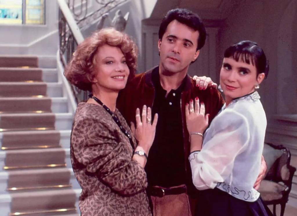 Cena de Rainha da Sucata com três personagens em um salão elegante, representando o universo de luxo e conflitos da novela que volta ao Vale a Pena Ver de Novo.