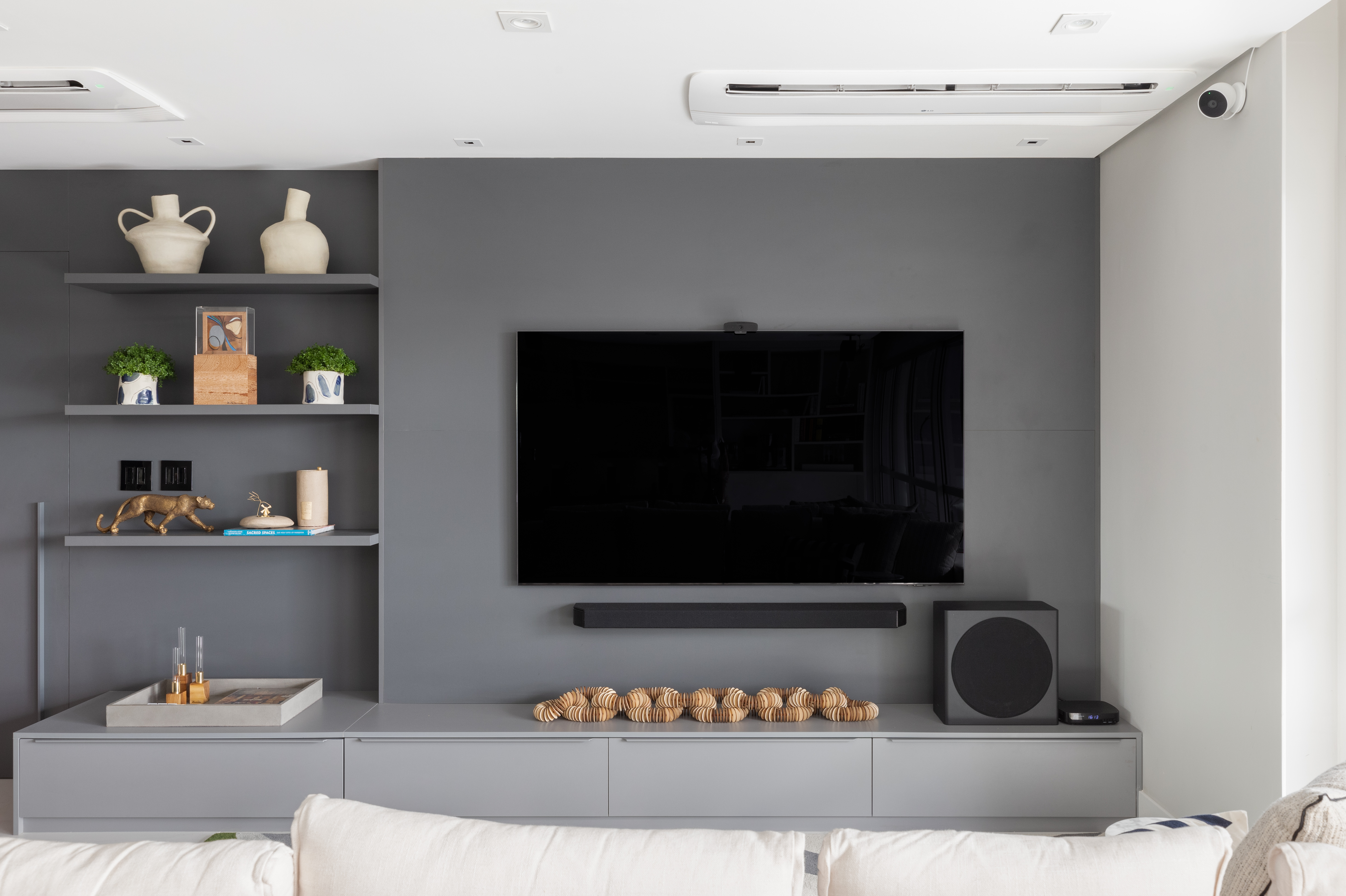 Painel de TV cinza com rack baixo e prateleiras laterais no mesmo tom, decoradas com vasos de cer&acirc;mica, livros, esculturas met&aacute;licas e pequenos arranjos verdes. A composi&ccedil;&atilde;o monocrom&aacute;tica destaca o televisor central e o subwoofer, criando um visual minimalista e elegante na sala de estar