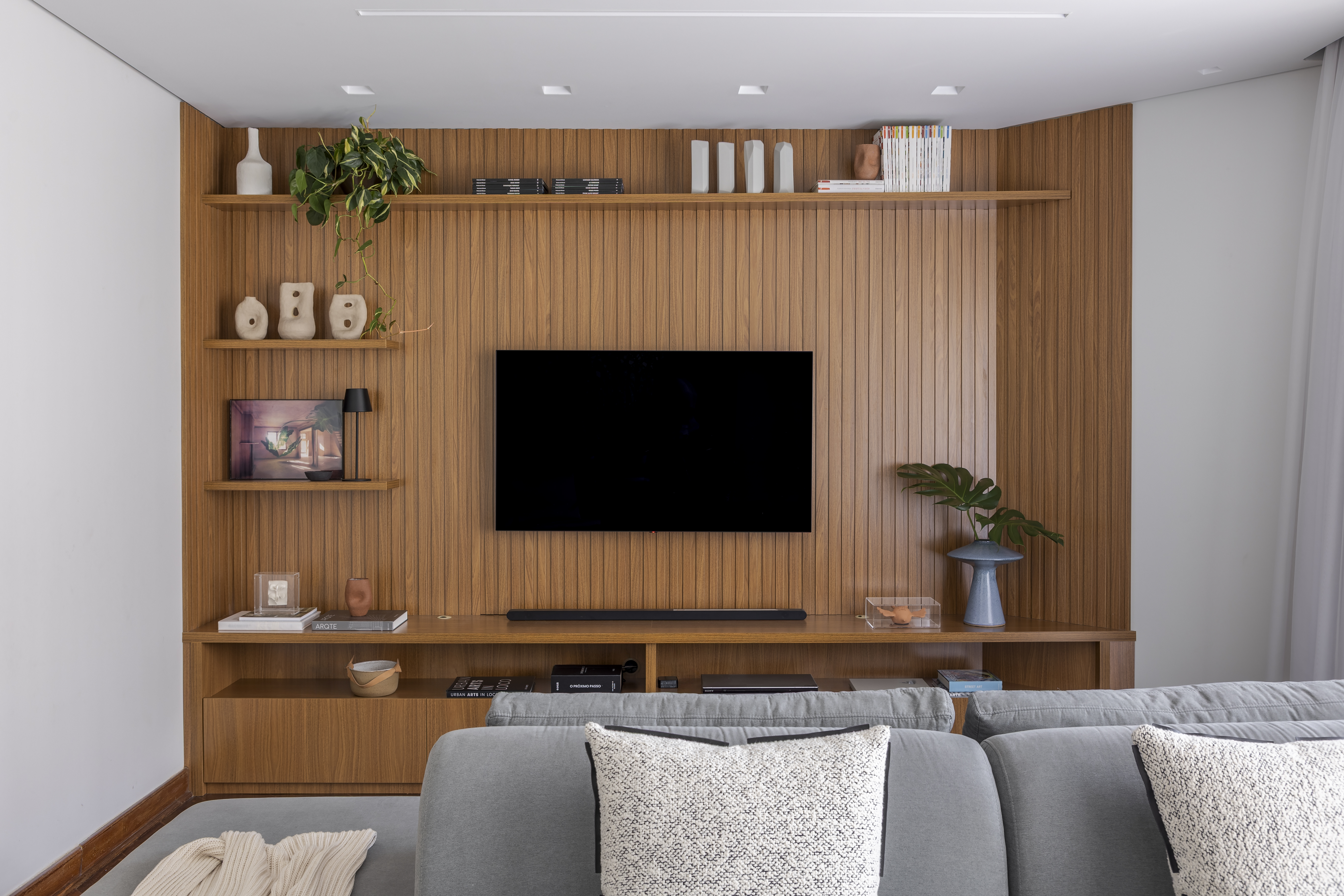 Painel ripado de madeira clara com prateleiras finas que exibem vasos escult&oacute;ricos, livros e plantas. O rack cont&iacute;nuo abriga aparelhos eletr&ocirc;nicos, caixas decorativas e adornos em vidro, enquanto o televisor preto cria contraste central. O ambiente tem est&eacute;tica contempor&acirc;nea com marcenaria limpa e bem iluminada