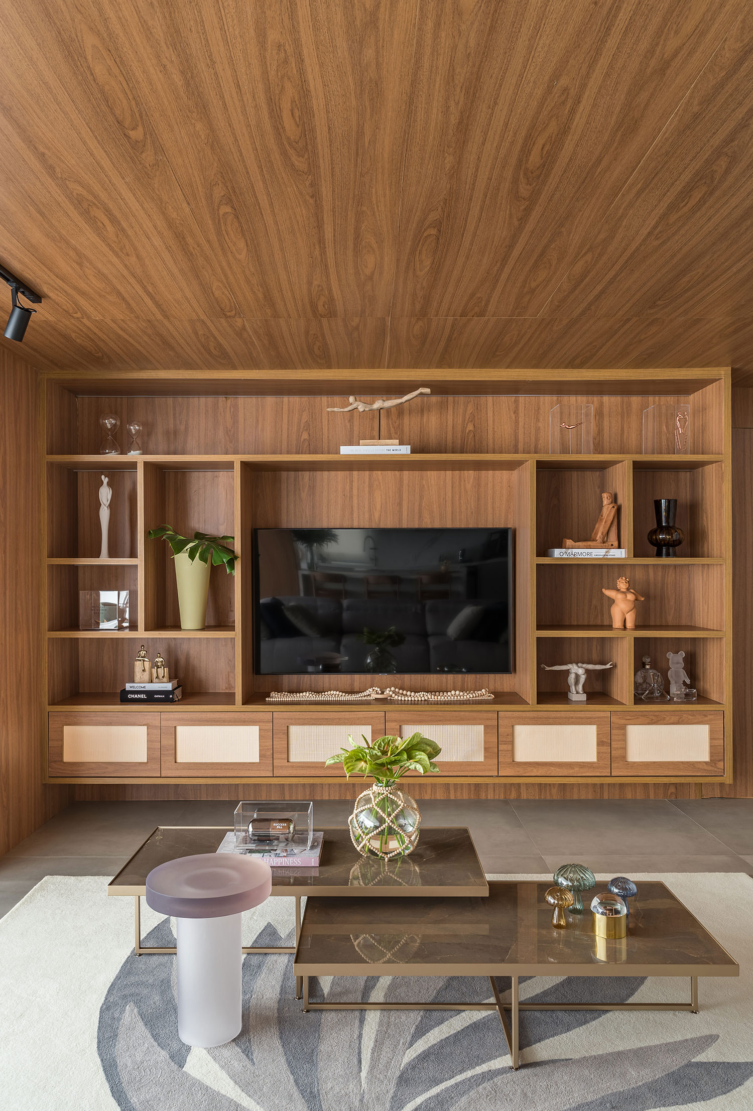 Estante e painel de TV totalmente amadeirados, com nichos abertos distribu&iacute;dos ao redor da tela e objetos decorativos como esculturas, vasos, pe&ccedil;as em vidro e livros. Uma mesa de centro dupla com tampo fum&ecirc; e vaso de plantas completa a sala integrada, marcada pela continuidade da madeira no teto e no mobili&aacute;rio