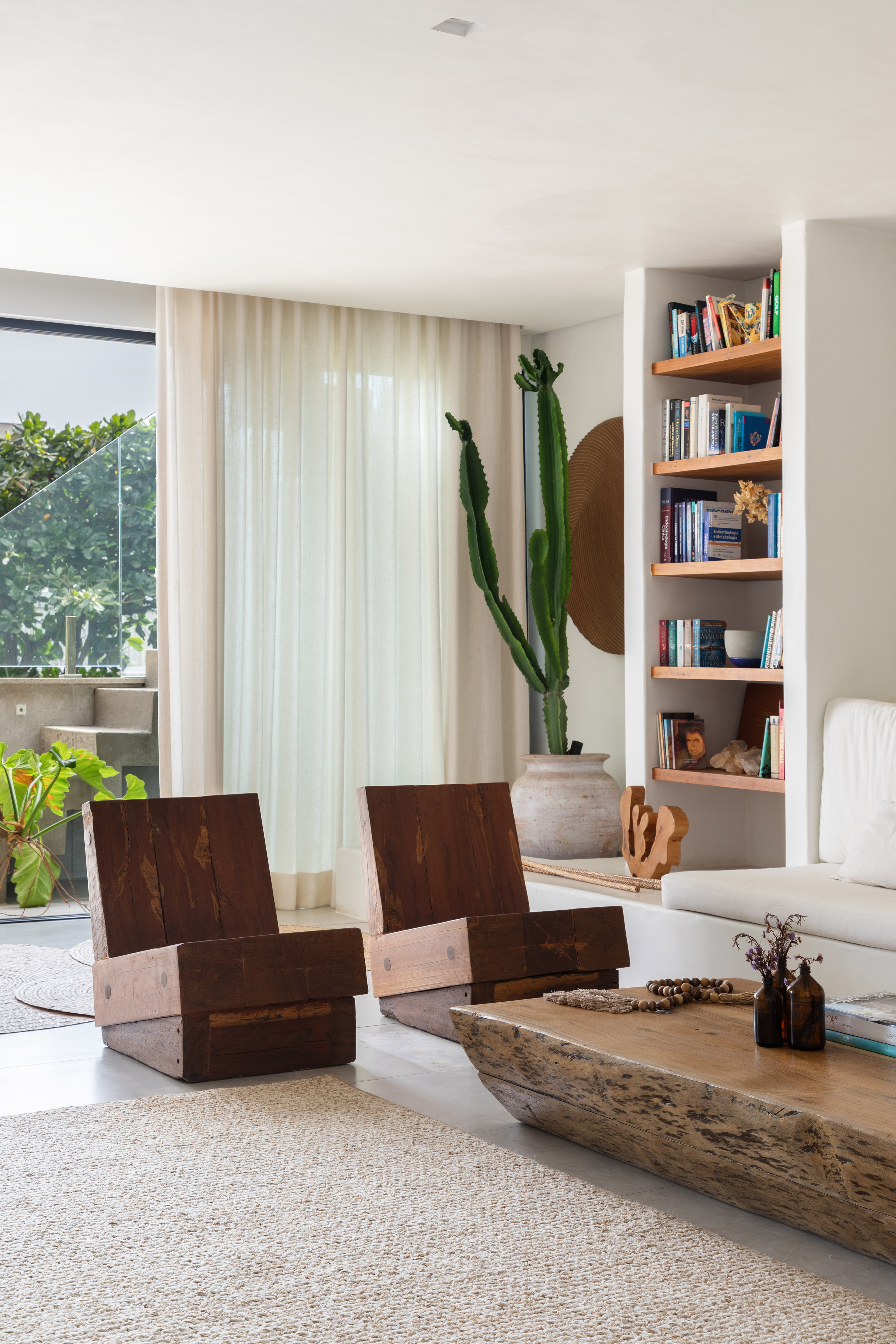 Foto de uma sala com decora&ccedil;&atilde;o predominantemente branca, com cactos, poltrona de madeira e estante de livros