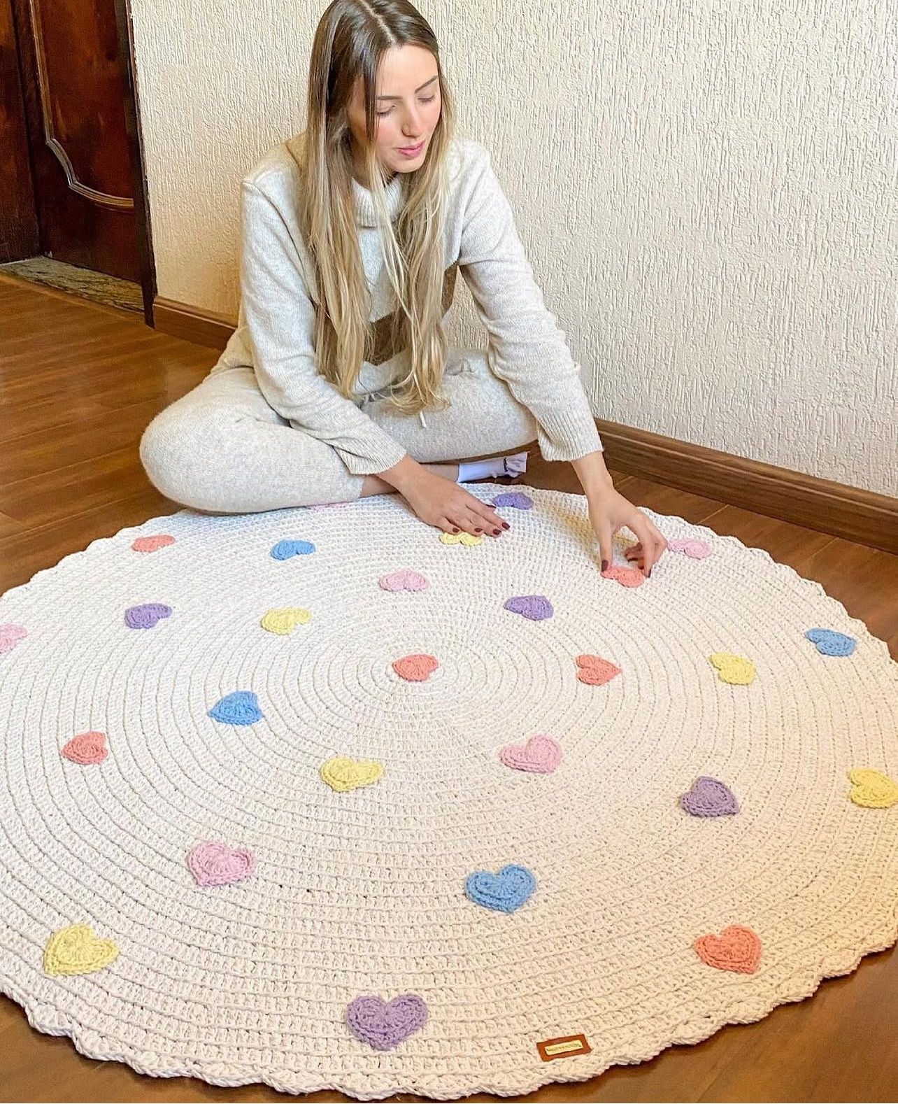 Foto de uma mulher loira sentada no ch&atilde;o. Ela toca um tapete infantil de croch&ecirc; branco com cora&ccedil;&otilde;es vermelhos