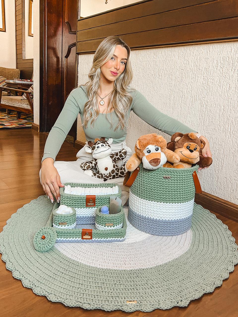 Foto de uma mulher loira sentada no ch&atilde;o. Ela toca um tapete infantil de croch&ecirc; verde, cinza e branco. H&aacute; ainda caixas com o mesmo material