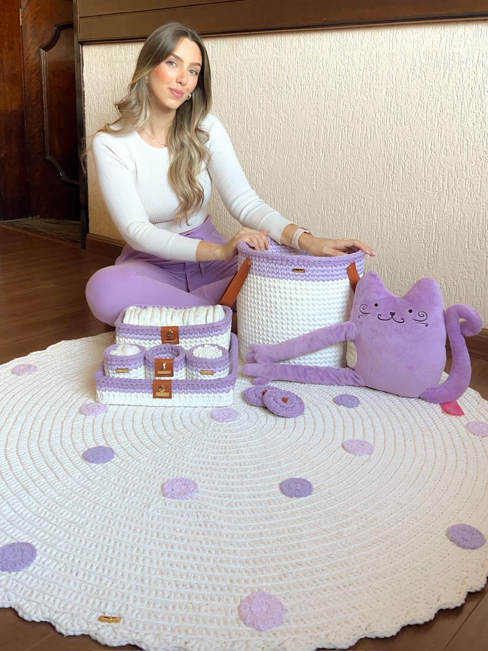 Foto de uma mulher loira sentada no ch&atilde;o. Ela toca um tapete infantil de croch&ecirc; roxo, lil&aacute;s e branco. Tamb&eacute;m h&aacute; caixas com a mesma decora&ccedil;&atilde;o