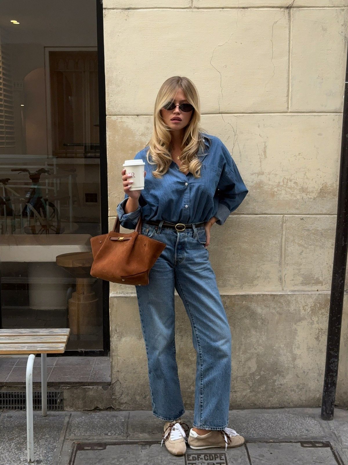 Foto de uma mulher loira usando um look composto por camisa jeans, cal&ccedil;a jeans, &oacute;culos de sol preto, bolsa marrom e t&ecirc;nis all star