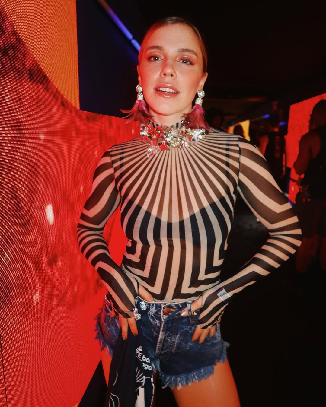 Foto de uma mulher loira usando um look composto por blusa transparente com detalhes branco e preto, colar brilhante e short jeans azul