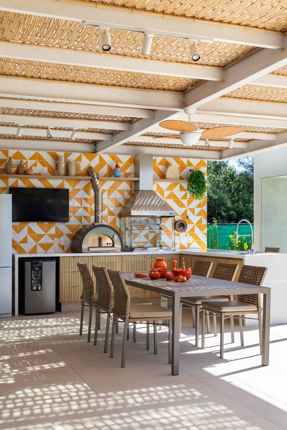 &Aacute;rea gourmet com azulejo laranja e branco, mesa com seis lugares retangular, churrasqueira e eletrodom&eacute;sticos em inox.
