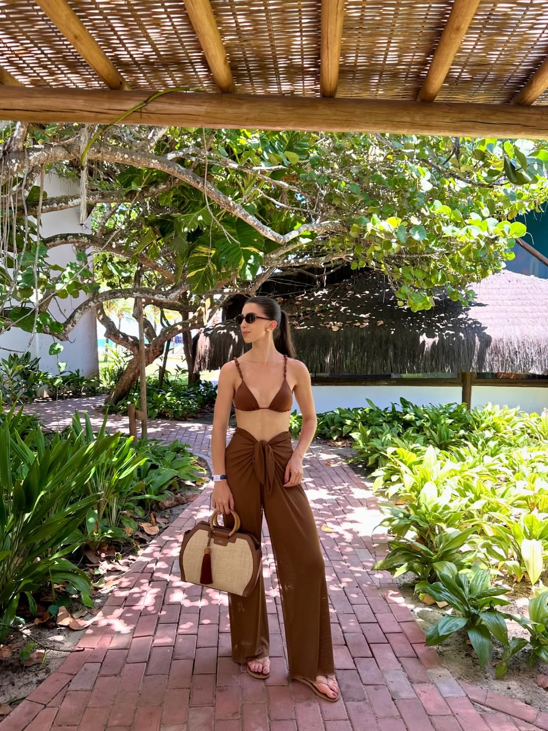 Foto de uma mulher morena usando um look composto por b&iacute;quini, cal&ccedil;a marrom, bolsa de palha, &oacute;culos de sol e sand&aacute;lia