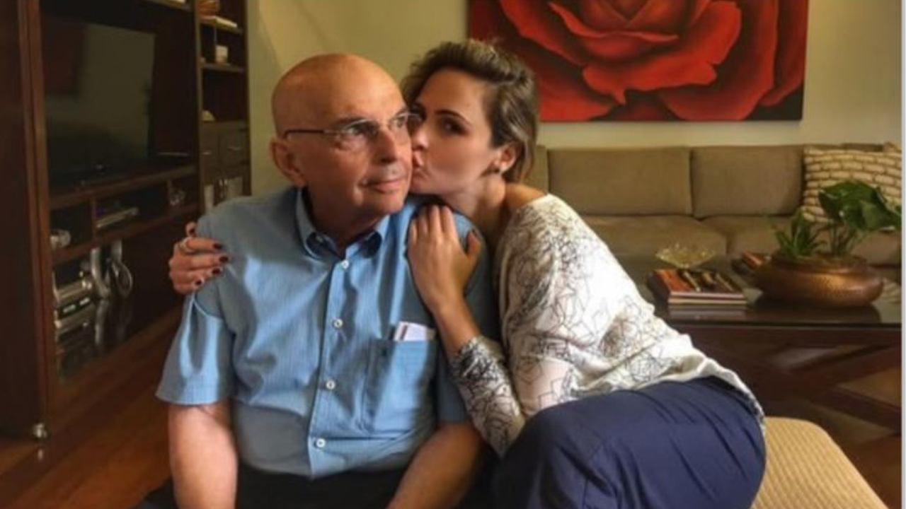 Um homem idoso, careca e usando óculos, está sentado no sofá com camisa azul clara. Ao lado dele, uma mulher mais jovem o abraça e dá um beijo em sua bochecha, com o braço apoiado em seus ombros. Ele olha para o lado com expressão serena, enquanto ela demonstra carinho