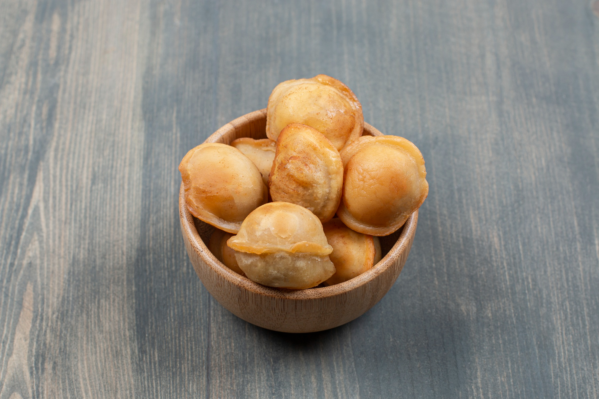 Uma tigela de madeira cheia de bolinhos de chuva dourados, com textura levemente crocante e apar&ecirc;ncia caseira, apoiada sobre uma mesa de madeira azul acinzentada.
