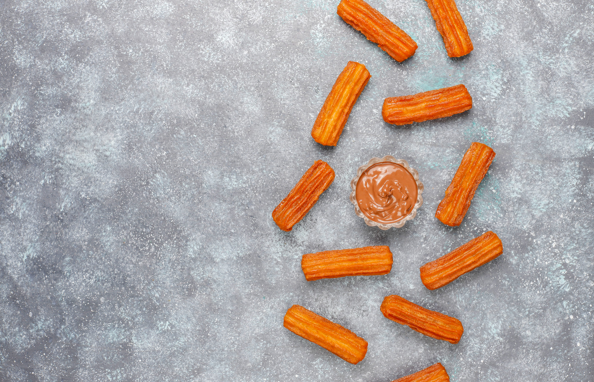 Mini churros dourados e crocantes dispostos em c&iacute;rculo ao redor de um potinho com doce de leite cremoso, sobre fundo cinza texturizado, com bastante espa&ccedil;o vazio na composi&ccedil;&atilde;o.