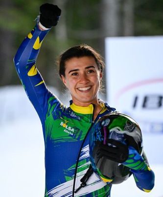 Nicole Silveira posa sorrindo ap&oacute;s a prova de skeleton, vestindo macac&atilde;o verde e azul do Brasil e segurando o capacete com uma das m&atilde;os, enquanto levanta o bra&ccedil;o em comemora&ccedil;&atilde;o