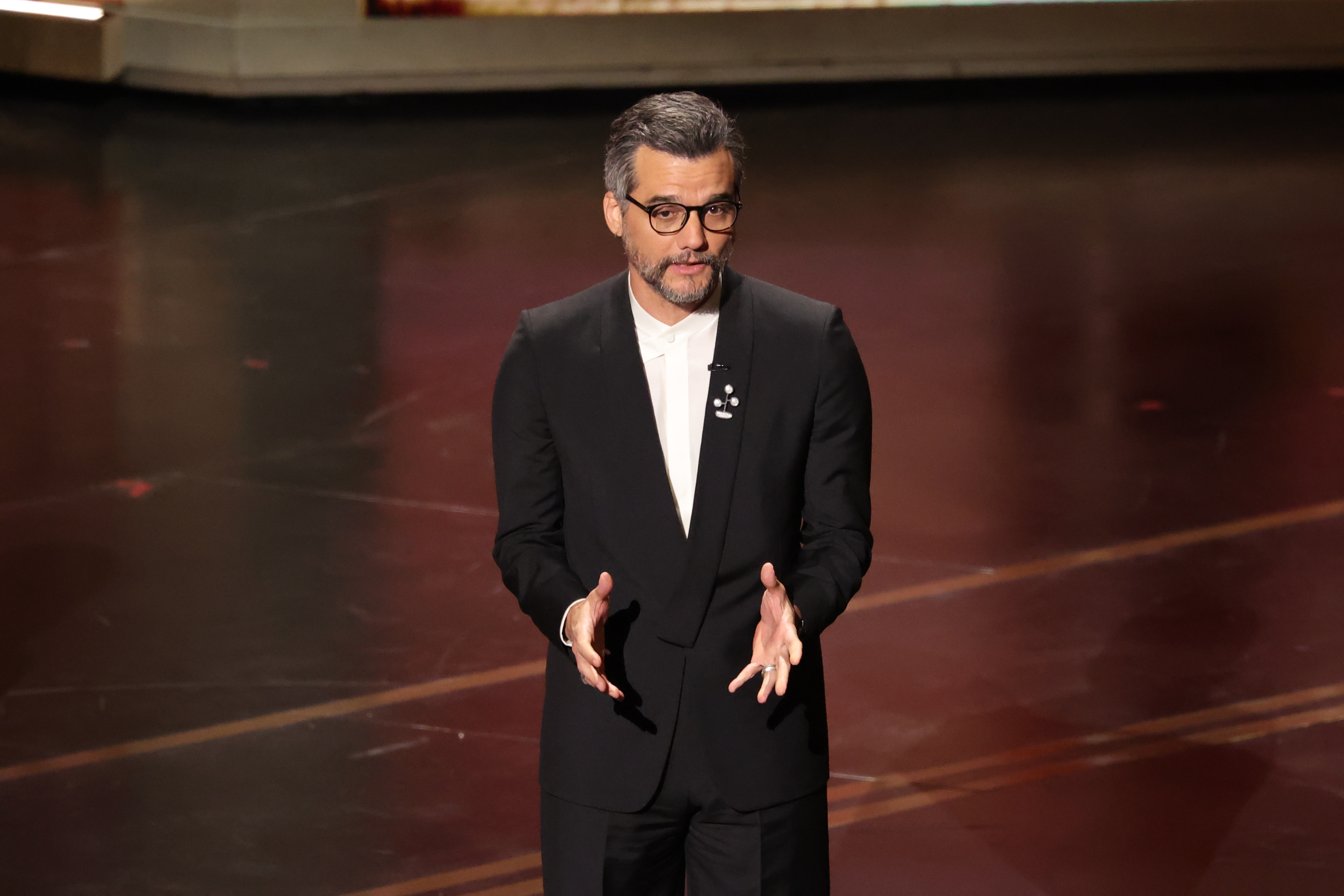 Wagner Moura aparece no palco do Oscar 2026 usando terno preto e camisa branca, com as mãos à frente do corpo durante discurso na cerimônia.