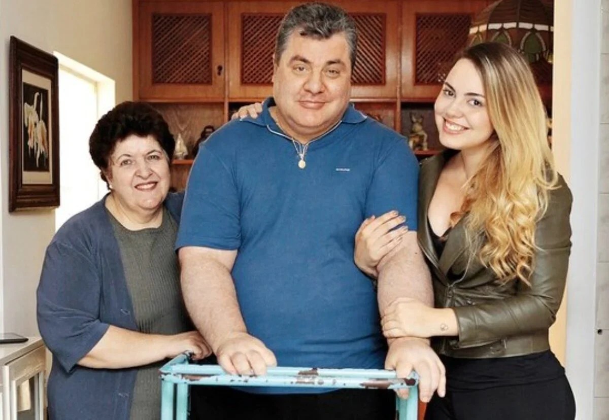 Gerson Brenner em casa, ao lado de duas mulheres da família, apoiado em uma estrutura de apoio, sorrindo para a câmera após período de recuperação