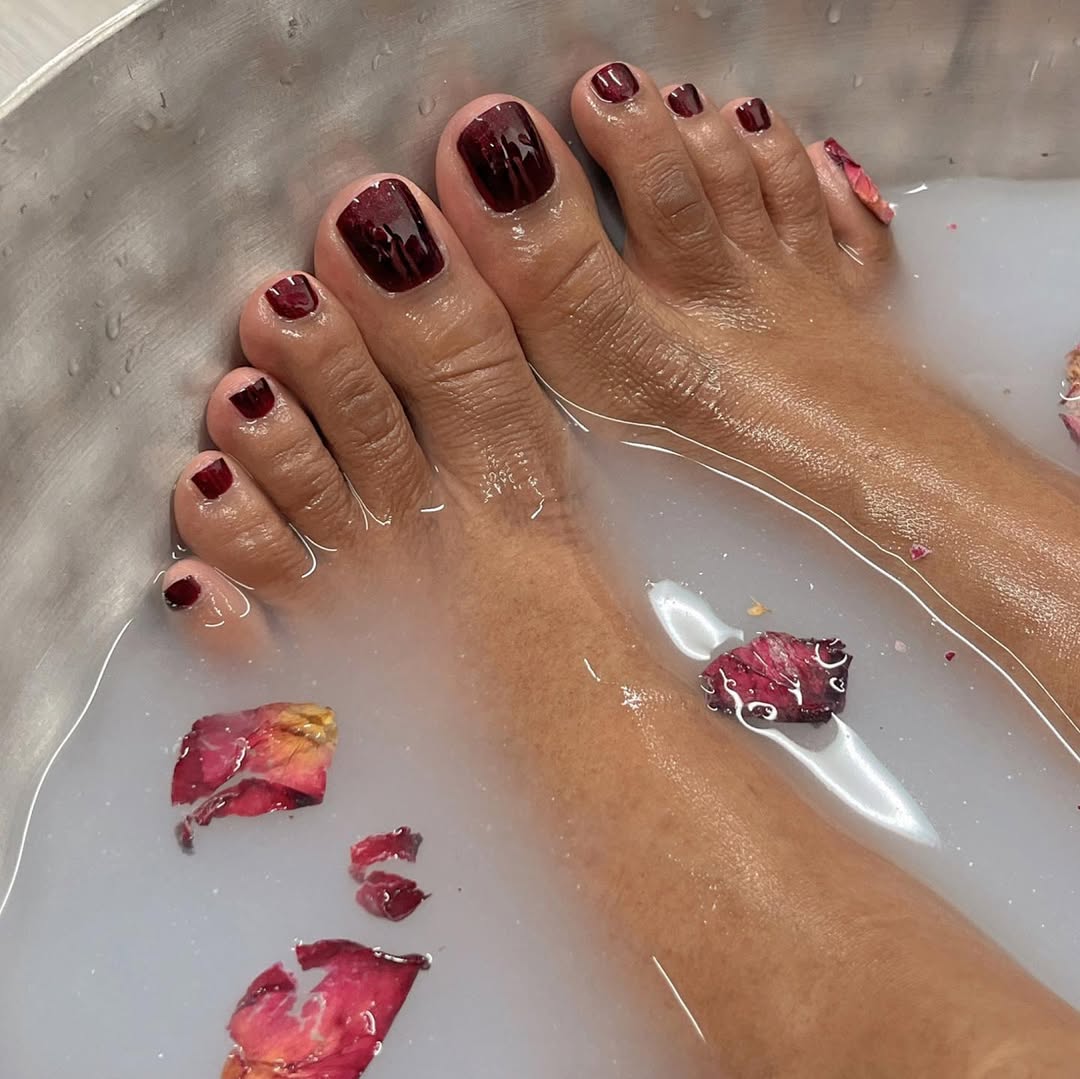 Foto de um p&eacute; de uma mulher negra com as unhas coloridas de vinho. Os p&eacute;s est&atilde;o numa bacia com flores e &aacute;gua