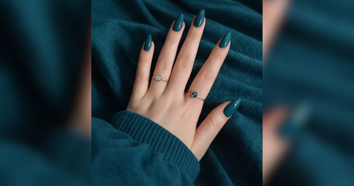 M&atilde;o feminina com unhas longas e pontiagudas pintadas de verde-escuro, usando dois an&eacute;is prateados com pedras claras, sobre um tecido verde-azulado