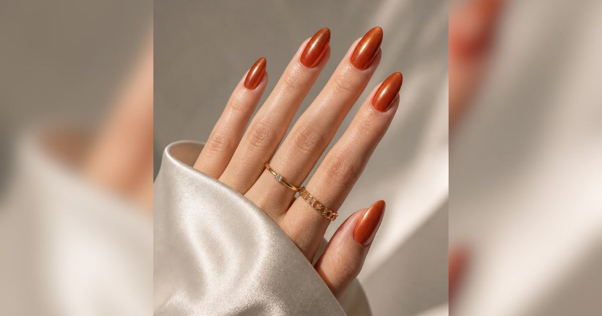 M&atilde;o feminina com unhas amendoadas pintadas de esmalte cobre met&aacute;lico, usando dois an&eacute;is dourados, um liso com pedra e outro de corrente, sobre tecido de seda bege
