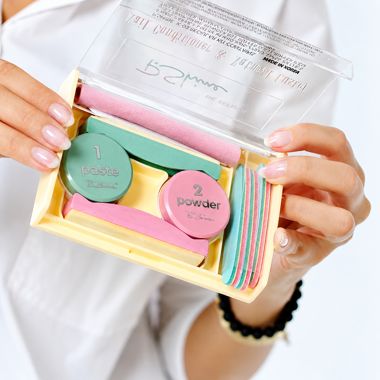 Mãos femininas seguram um kit de manicure amarelo pastel aberto, revelando potes de pasta verde e pó rosa, lixas e polidores de unhas em tons de rosa, verde e amarelo