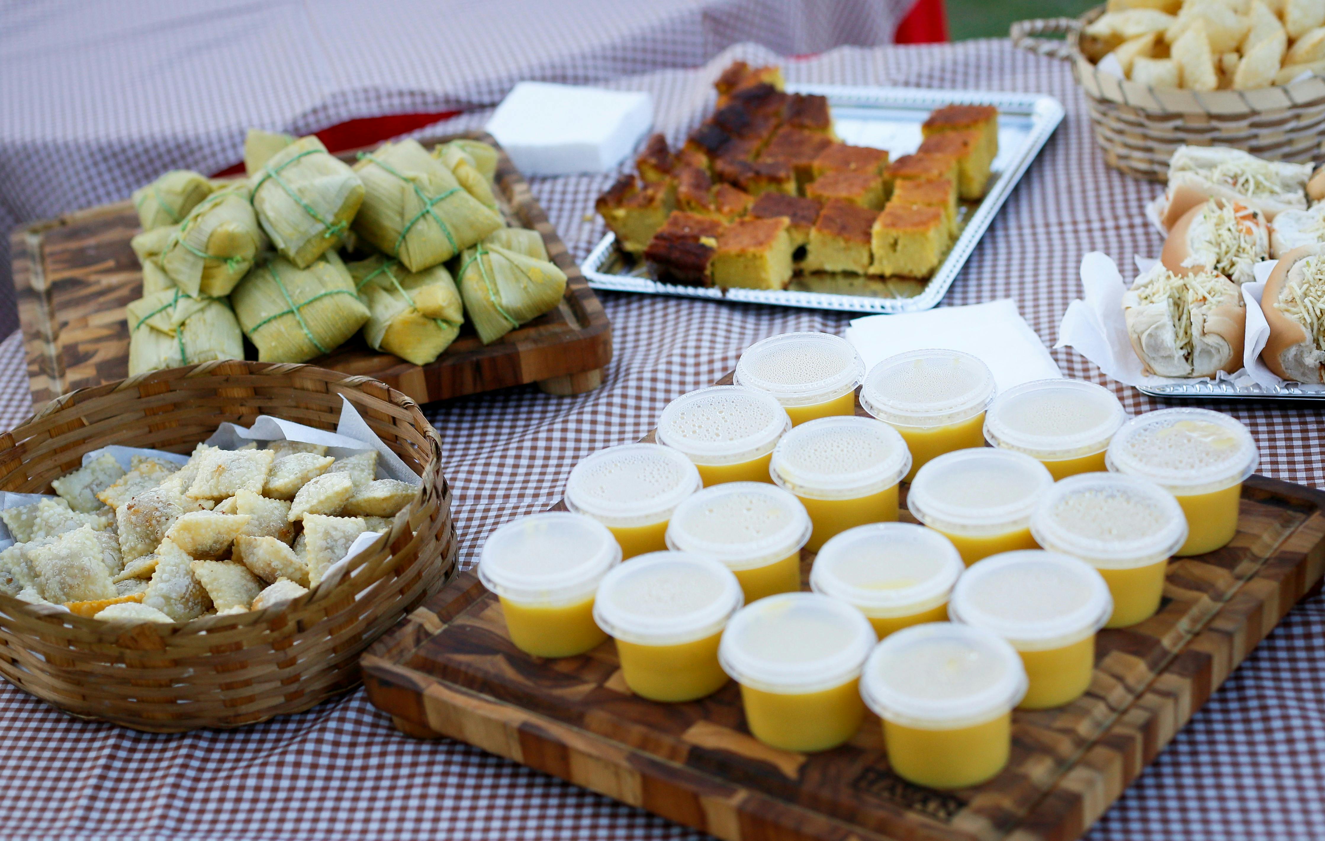 Mesa com toalha xadrez marrom e branca, exibindo comidas t&iacute;picas: pamonhas, bolo de milho, mini-past&eacute;is, curau em potes e p&atilde;es com recheio cremoso