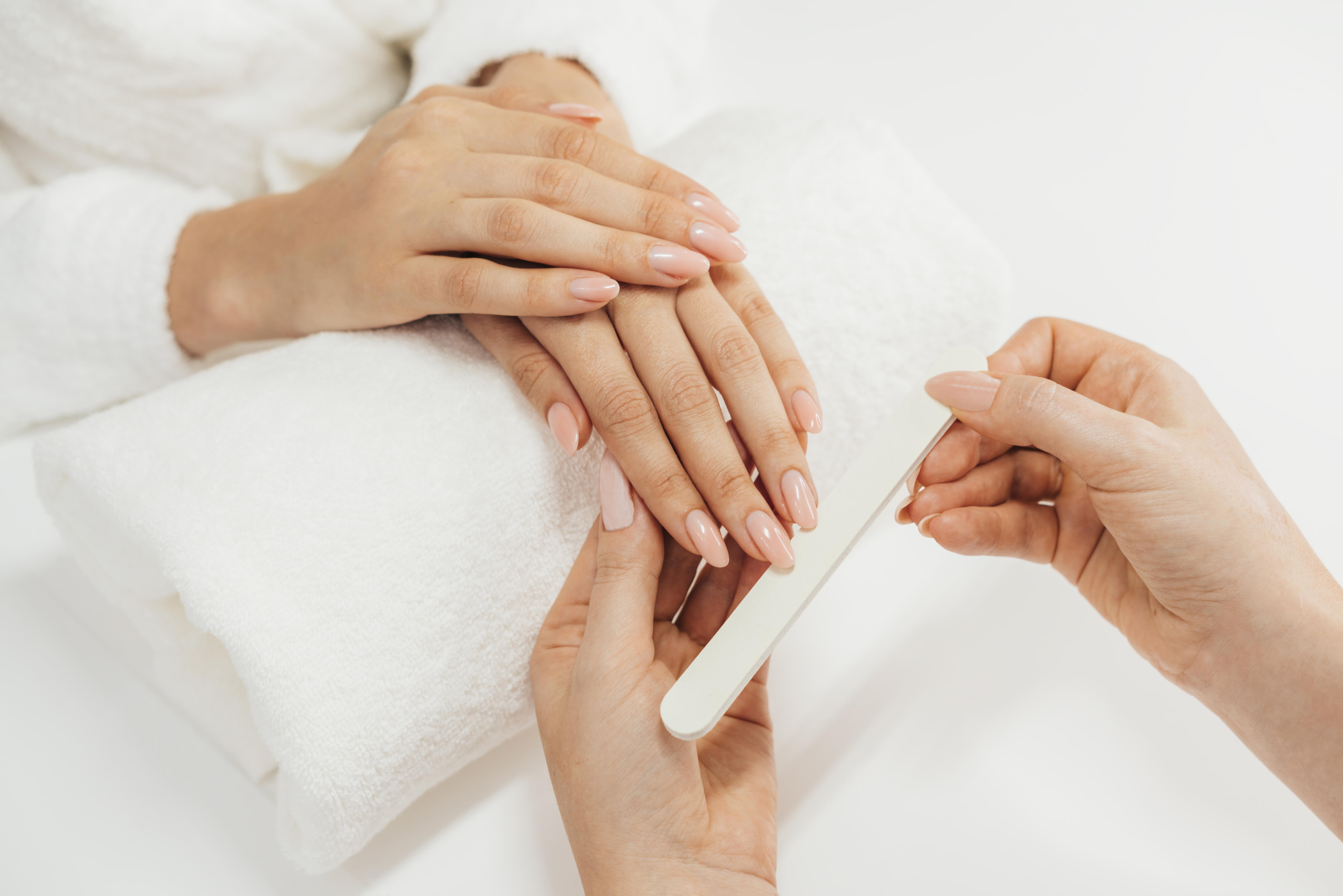 Duas m&atilde;os femininas recebendo manicure, com unhas pintadas em tom nude, sobre uma toalha branca e uma lixa de unhas sendo usada