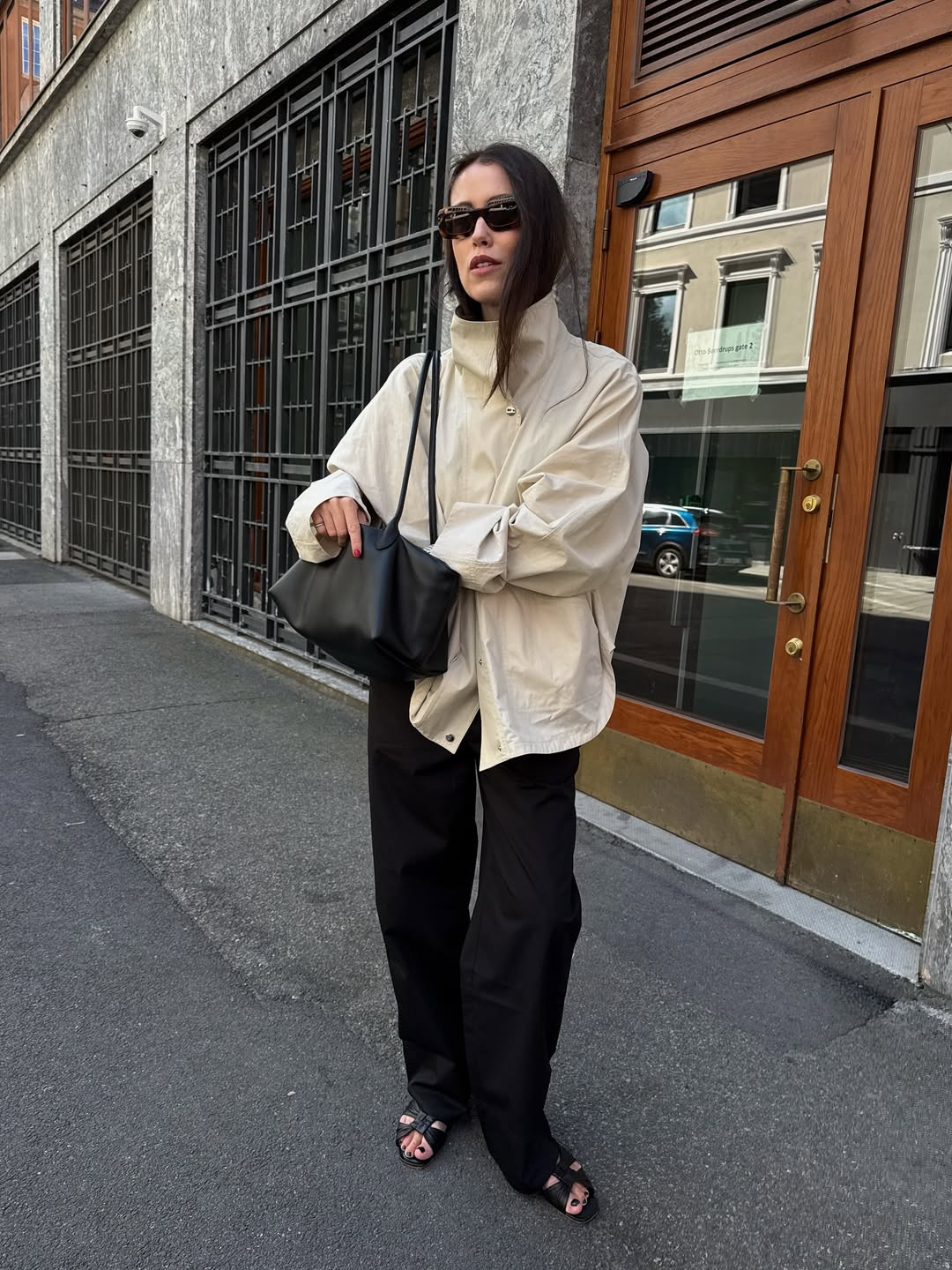 Foto de uma mulher branca usando um look composto por jaqueta parka bege, cal&ccedil;a preta, bolsa preta e salto