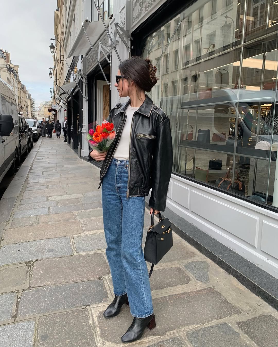 Foto de uma mulher branca, com cabelo preto, usando um look composto por jaqueta de couro, cal&ccedil;a jeans reta, bolsa preta e bota