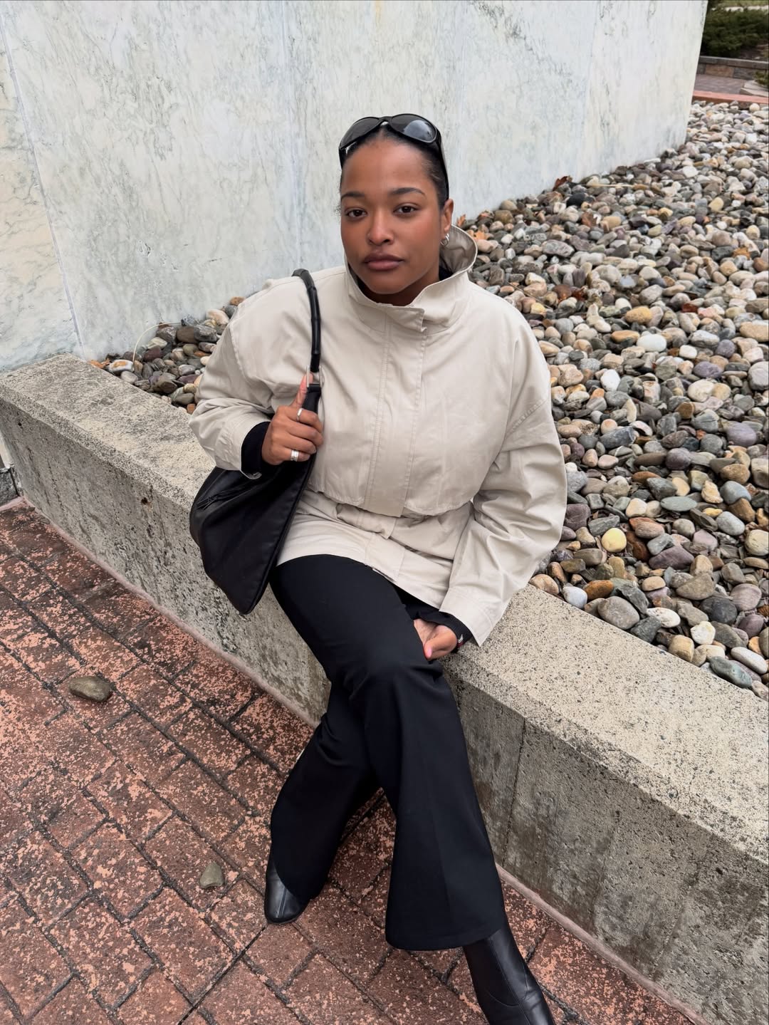 Foto de uma mulher negra usando um look composto por jaqueta parka bege, bolsa preta, cal&ccedil;a preta e bota de couro