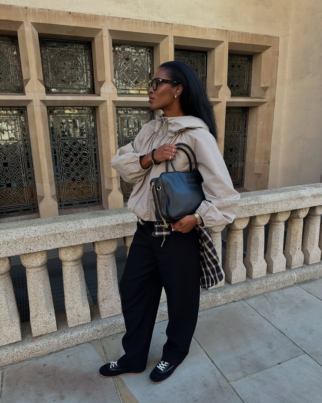 Foto de uma mulher negra usando um look composto por parka bege, cal&ccedil;a preta, bolsa de couro e sapatilha