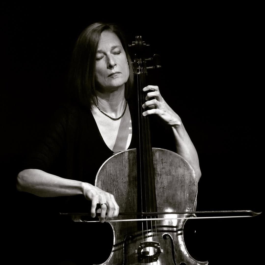 Em uma fotografia em preto e branco, a violoncelista alem&atilde; Anja Lechner toca violoncelo com os olhos fechados em um palco escuro. A imagem destaca a intensidade emocional da musicista e sua forte conex&atilde;o com a m&uacute;sica cl&aacute;ssica e experimental.