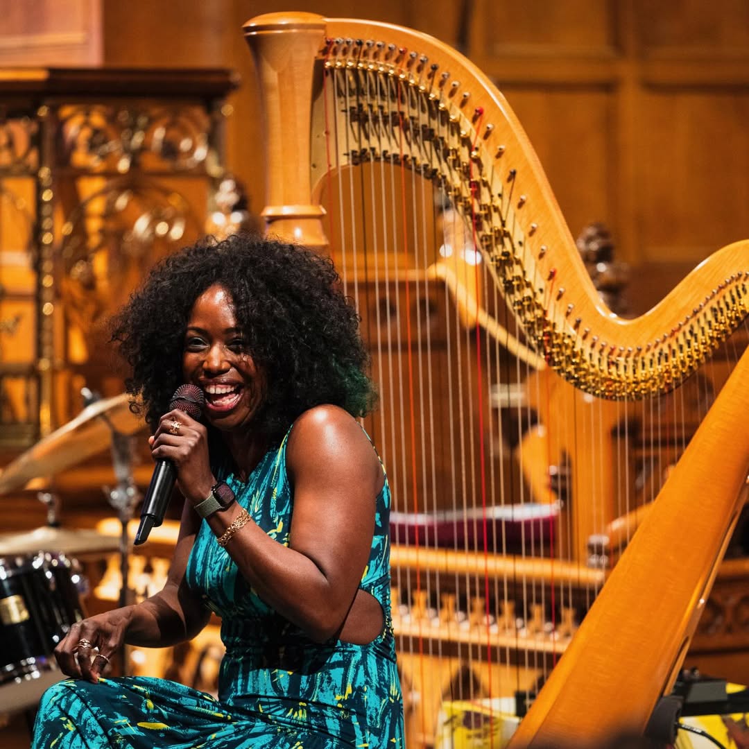 A harpista Brandee Younger se apresenta em um palco iluminado com tons dourados, segurando um microfone ao lado de uma harpa cl&aacute;ssica. Usando vestido estampado em azul e verde, a artista transmite a energia do jazz contempor&acirc;neo ao unir sofistica&ccedil;&atilde;o, improviso e refer&ecirc;ncias de R&B e hip-hop.