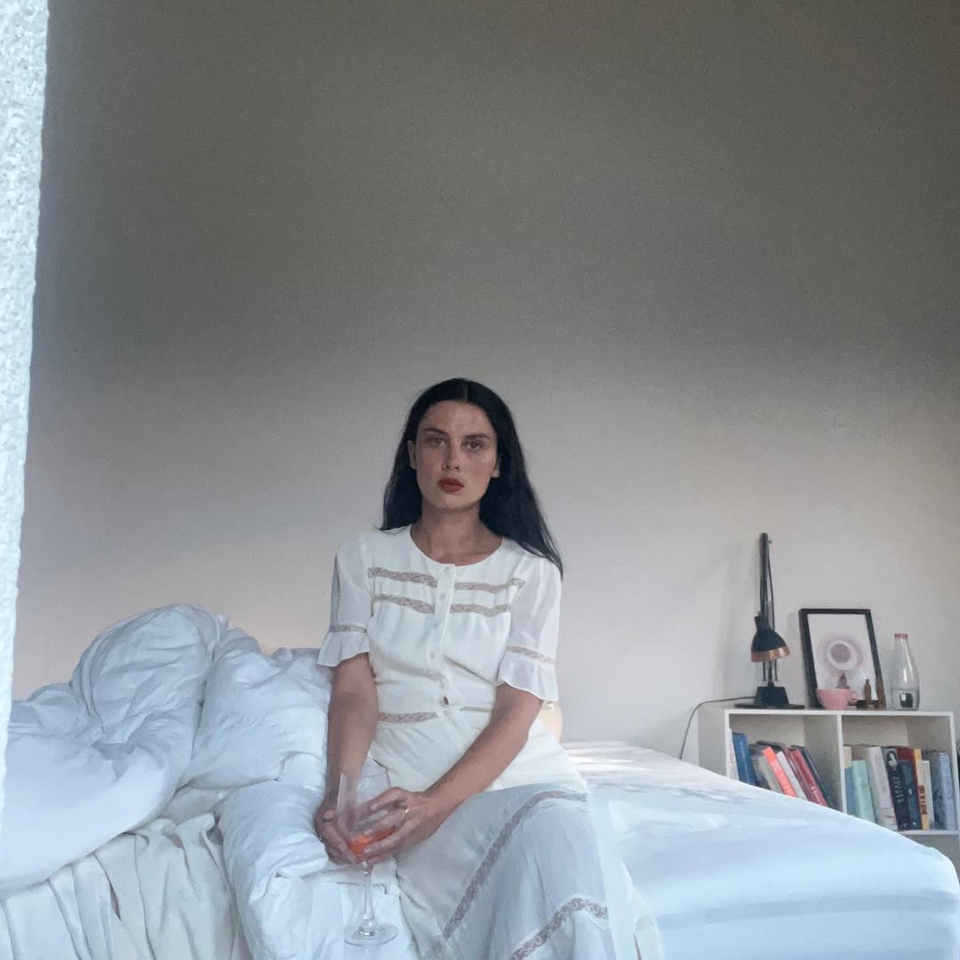 Ellie Rowsell posa sentada em um quarto minimalista usando vestido branco delicado enquanto segura uma ta&ccedil;a rosada. A imagem suave e contemplativa combina com o universo experimental da violoncelista e compositora, conhecida por misturar cello, voz e atmosferas sonoras et&eacute;reas.