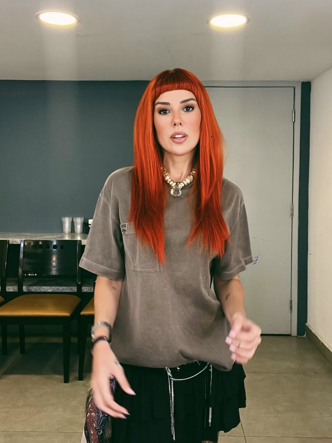 A DJ e produtora Aline Rocha aparece em camarim usando cabelo vermelho vibrante, camiseta oversized e acess&oacute;rios metalizados. O visual moderno e urbano conversa com a trajet&oacute;ria da artista brasileira na cena da house music e dos grandes festivais internacionais.