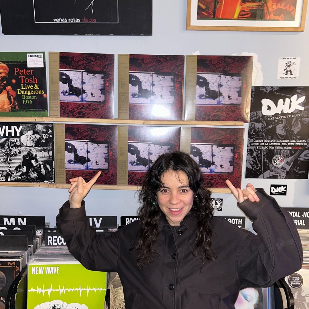 Mabe Fratti posa em uma loja de discos cercada por capas de vinil e p&ocirc;steres de bandas alternativas. Vestindo casaco escuro e fazendo gesto divertido para a c&acirc;mera, a vocalista do Wolf Alice refor&ccedil;a a est&eacute;tica indie rock e underground brit&acirc;nica.