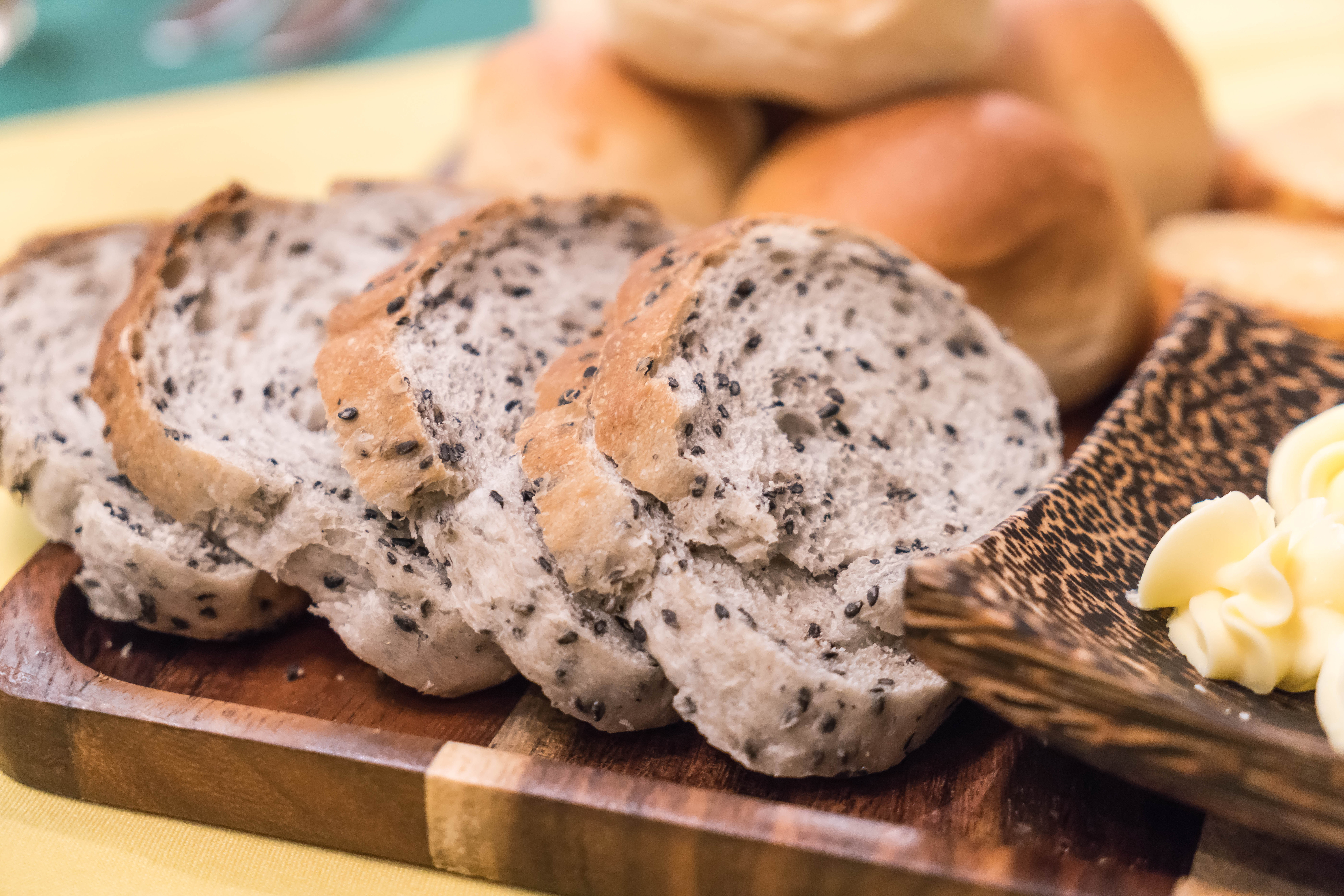 Close em fatias de p&atilde;o multigr&atilde;os com chia e sementes espalhadas pela massa. O p&atilde;o aparece servido sobre uma t&aacute;bua de madeira, acompanhado de manteiga e outros p&atilde;es ao fundo