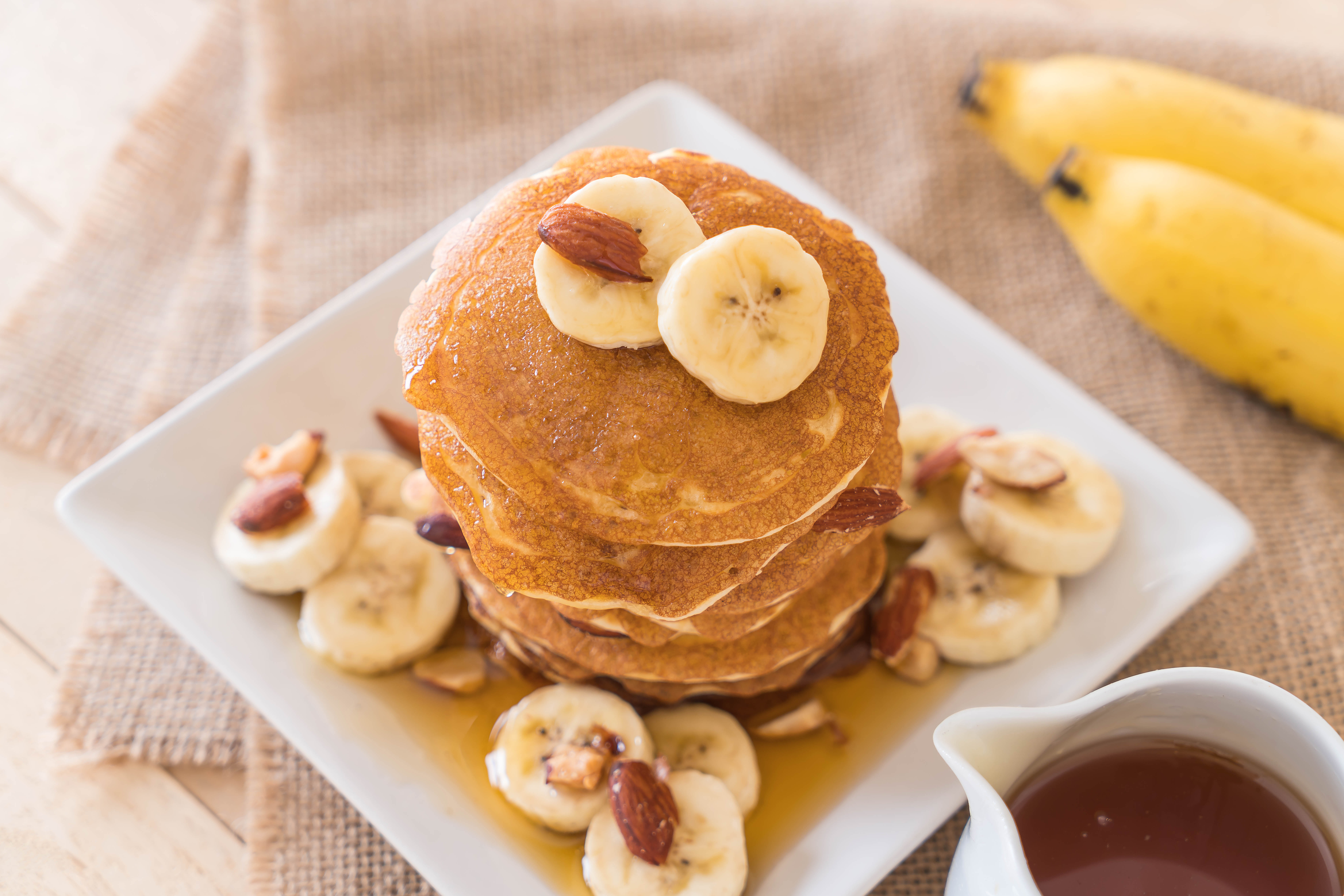 A fotografia destaca uma torre de panquecas douradas cobertas com banana, am&ecirc;ndoas e mel